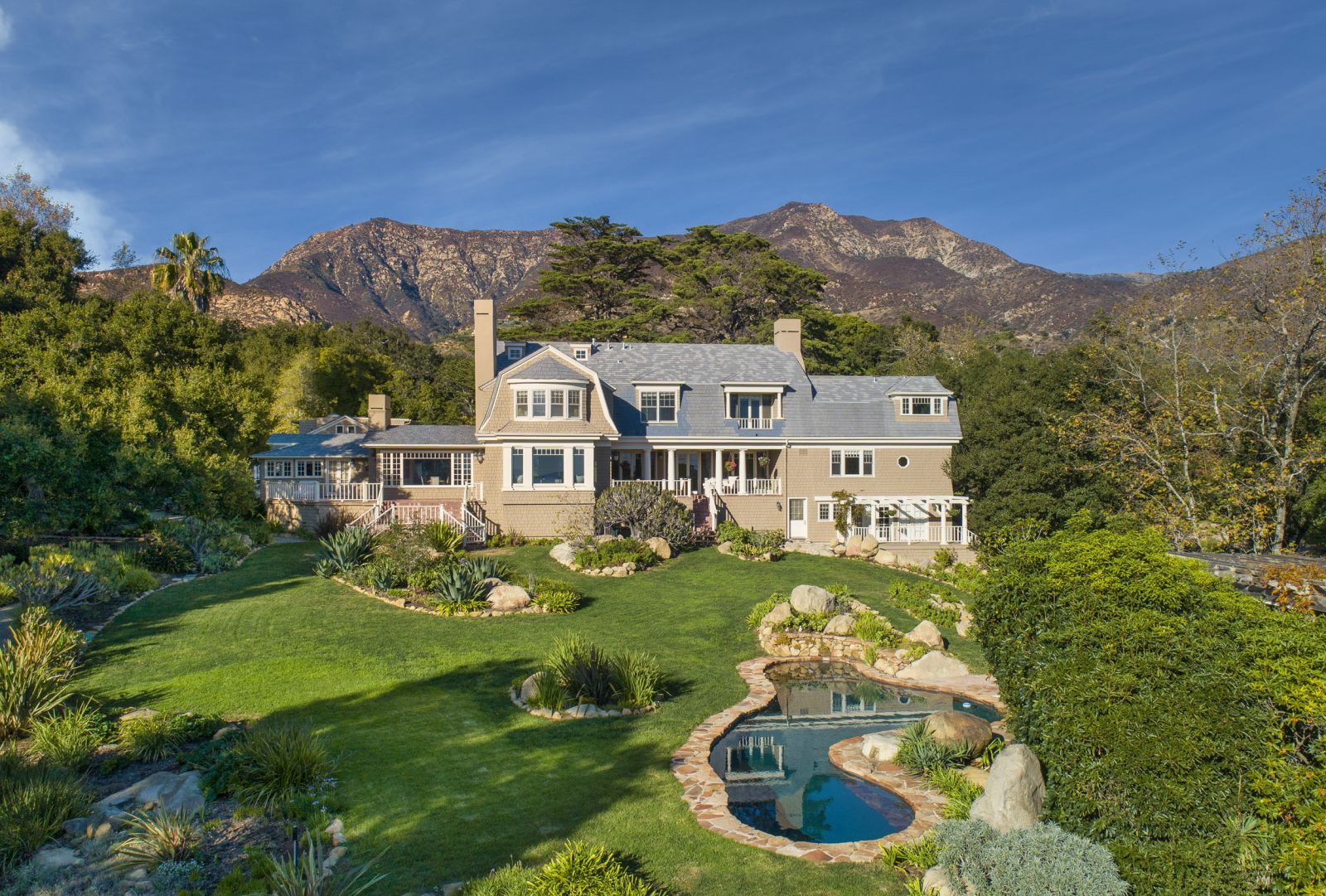 The large backyard of a luxury Cape Cpod-style Montecito home, with a lush lawn, and a pool framed by mountains.