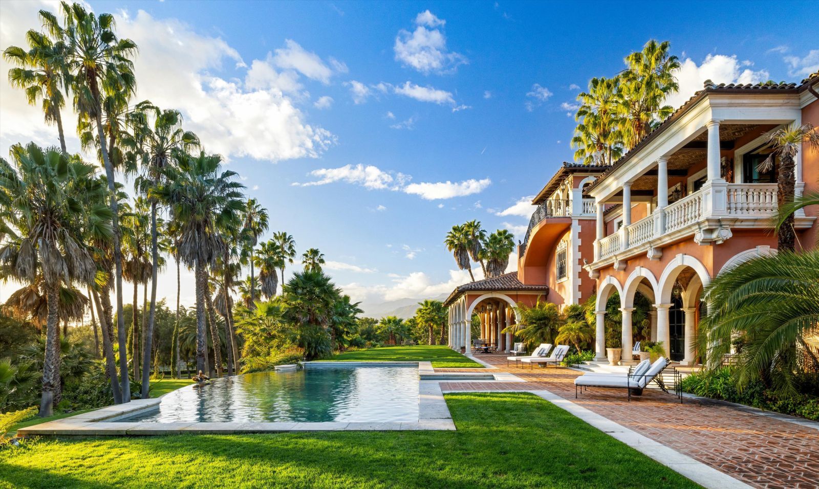 Ochre-colored rear facade of a Spanish Colonial Revival mansion in Montecito with a loggia over a veranda, a sparkling pool, palm trees, and brick terrace.