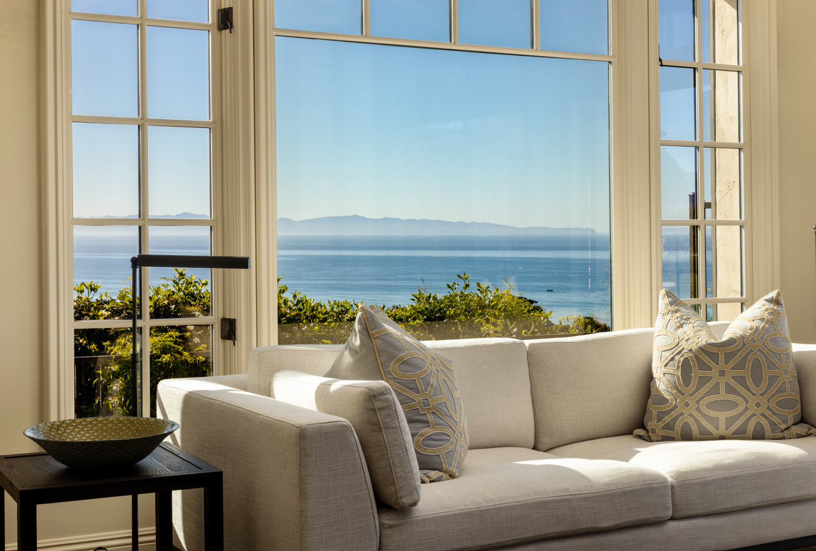 A couch with pillows sits in front of a floor to ceiling window with a view of the ocean.