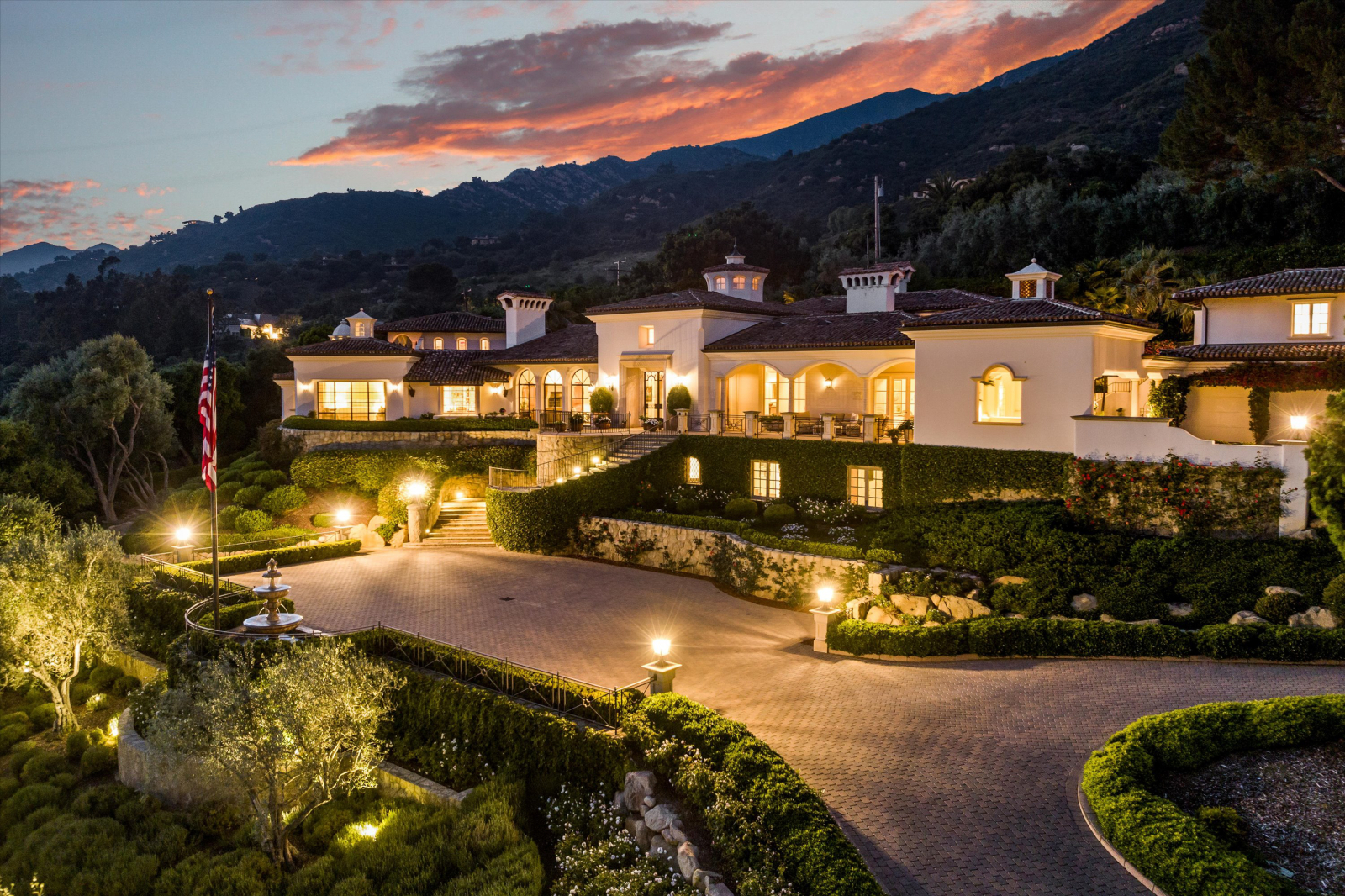 Luxury Montecito estate at sunset featuring Mediterranean-inspired architecture, illuminated terraces, manicured gardens, and a circular driveway set against the Santa Ynez Mountains.