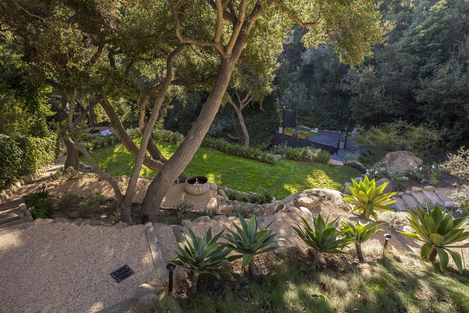 A meandering pathway through small boulders, native plants and trees, down to a green lawn below.