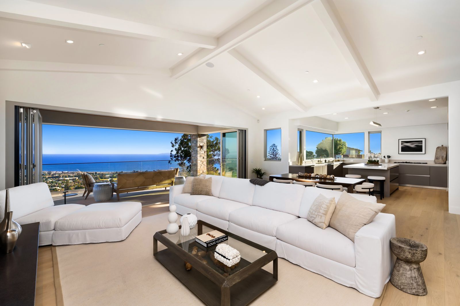 A bright and airy luxury living room with plush white furniture, a white open-beam ceiling, and a wall of glass offering an ocean view.