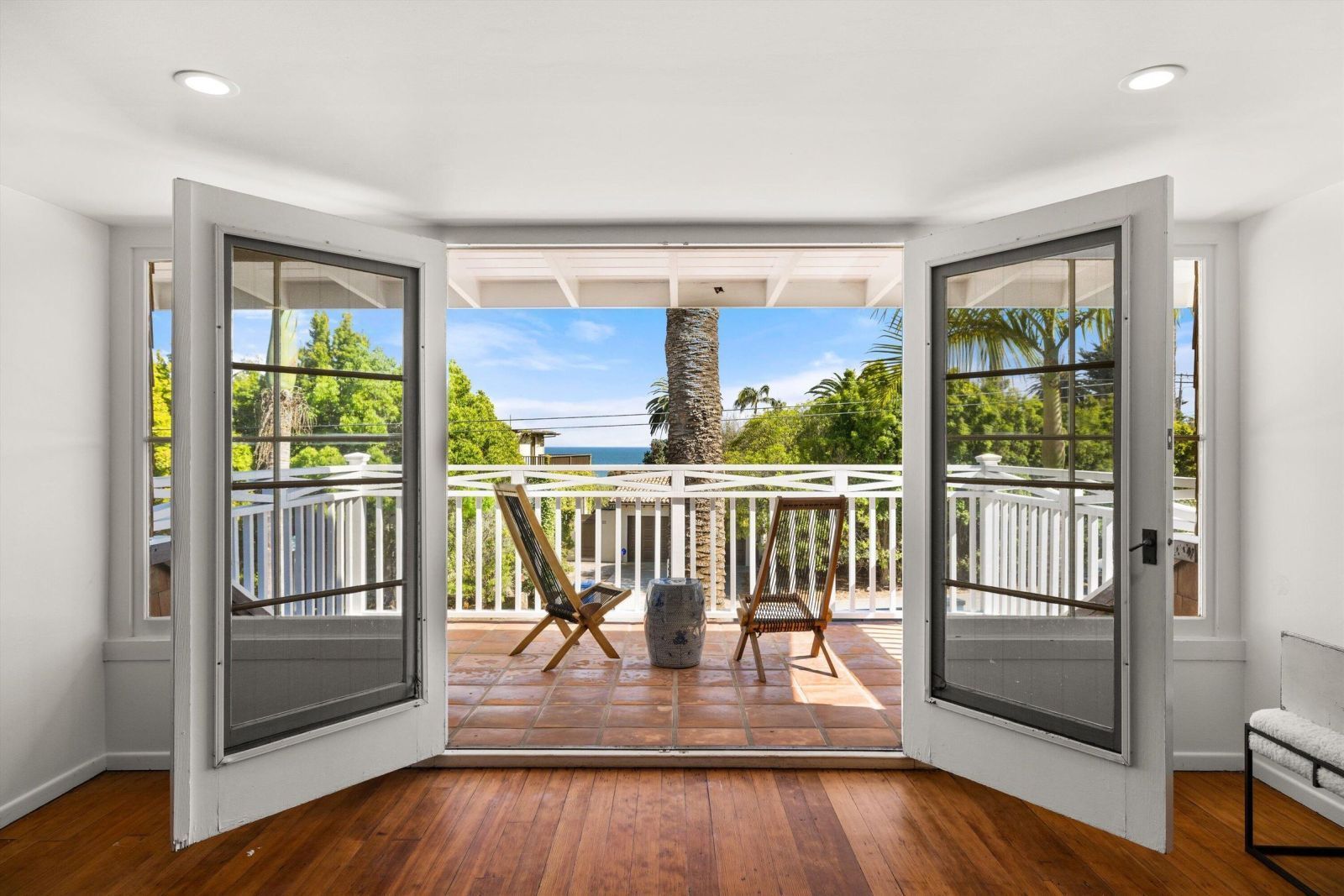 Doors open to the tereace of a Miramar Beach Estate, with a white picket fence and two chairs and a table showcasing mature tropical gardens and an ocean view