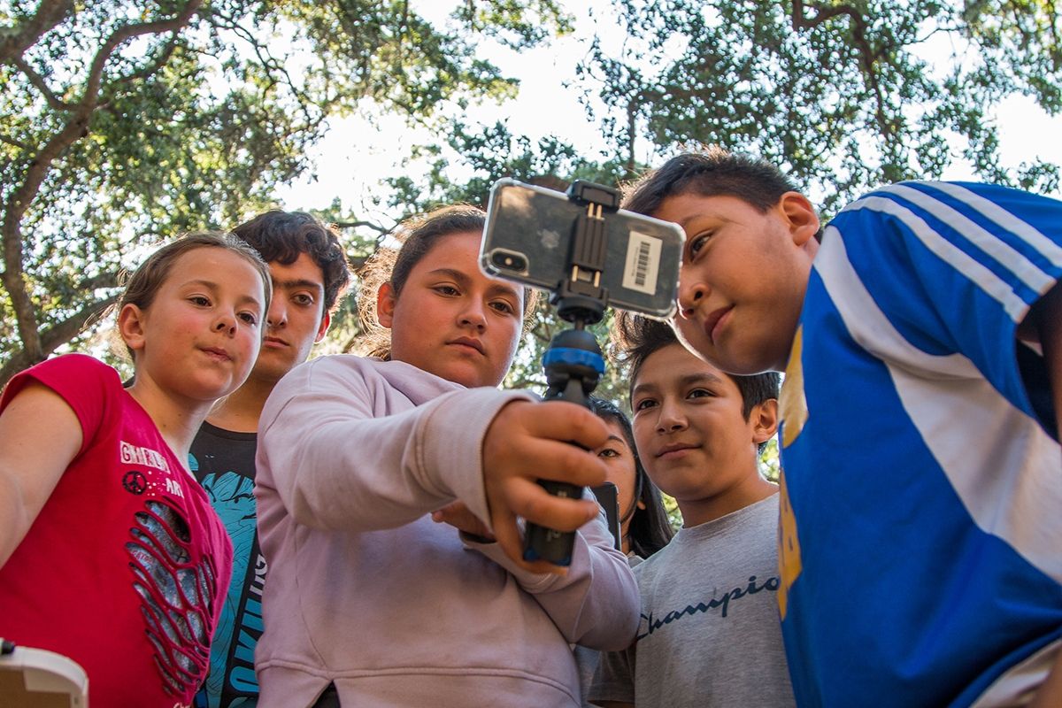 Five pre-teens outside in tall trees, huddled and ready to use a selfie stick and phone