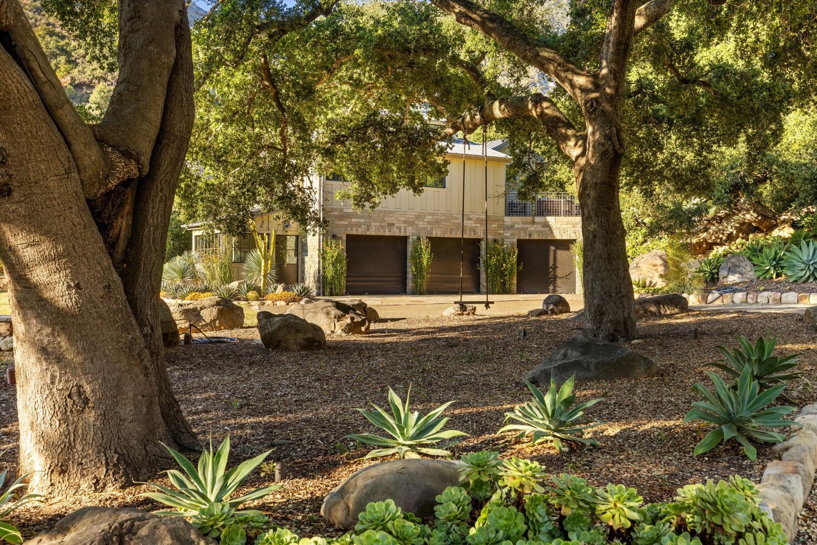 The backyard of a home with a pair of mature trees and a swing in the foreground.