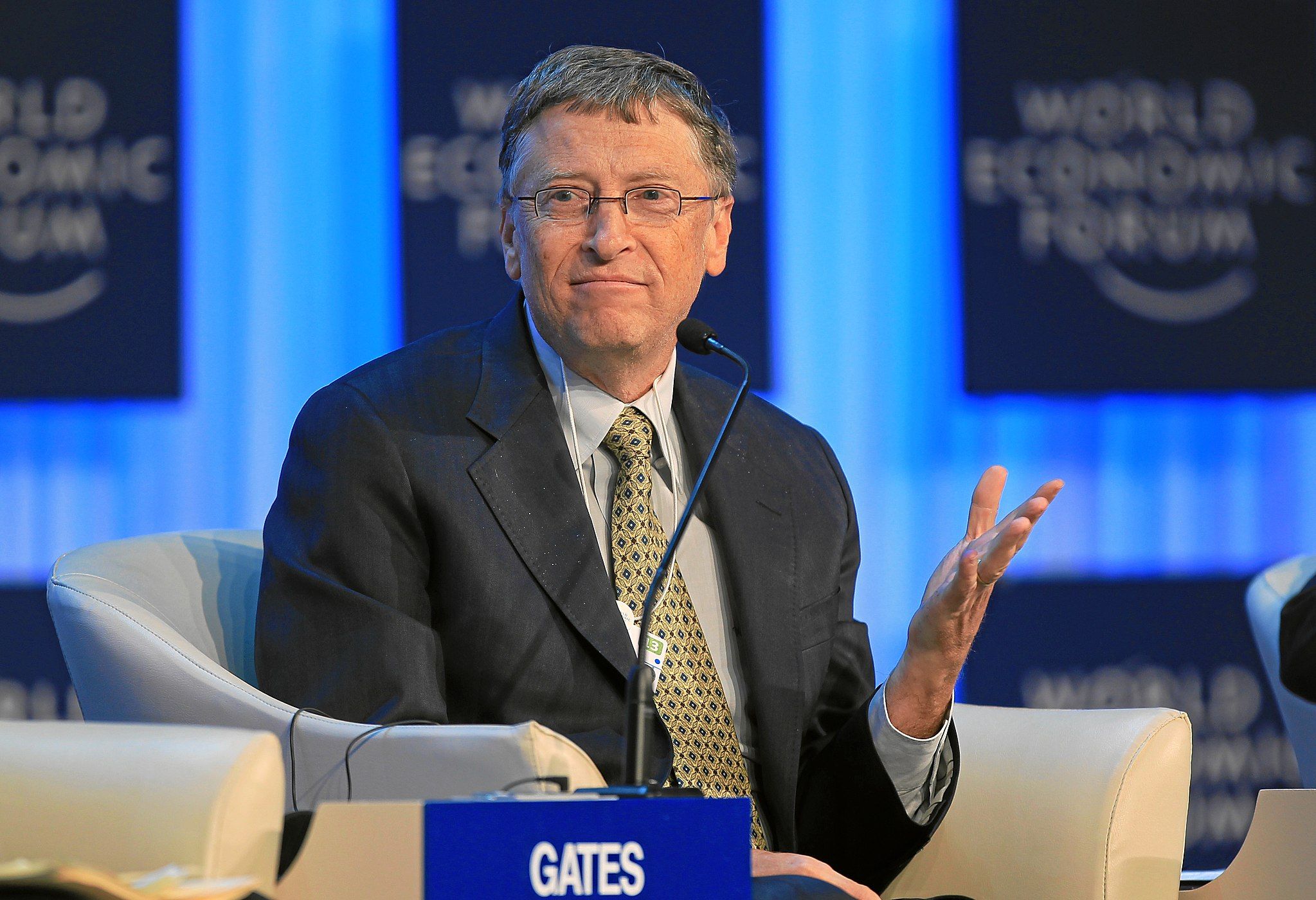 B ill Gates in a suit and tie sitting in front of a microphone.