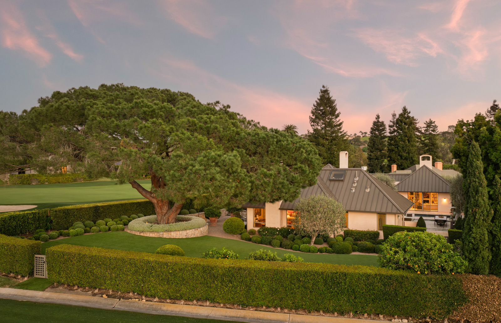 Beautifully landscaped estate featuring a single-story home with a metal roof, surrounded by manicured hedges, sculpted greenery, and a large mature tree, with a golf course visible nearby at sunset.