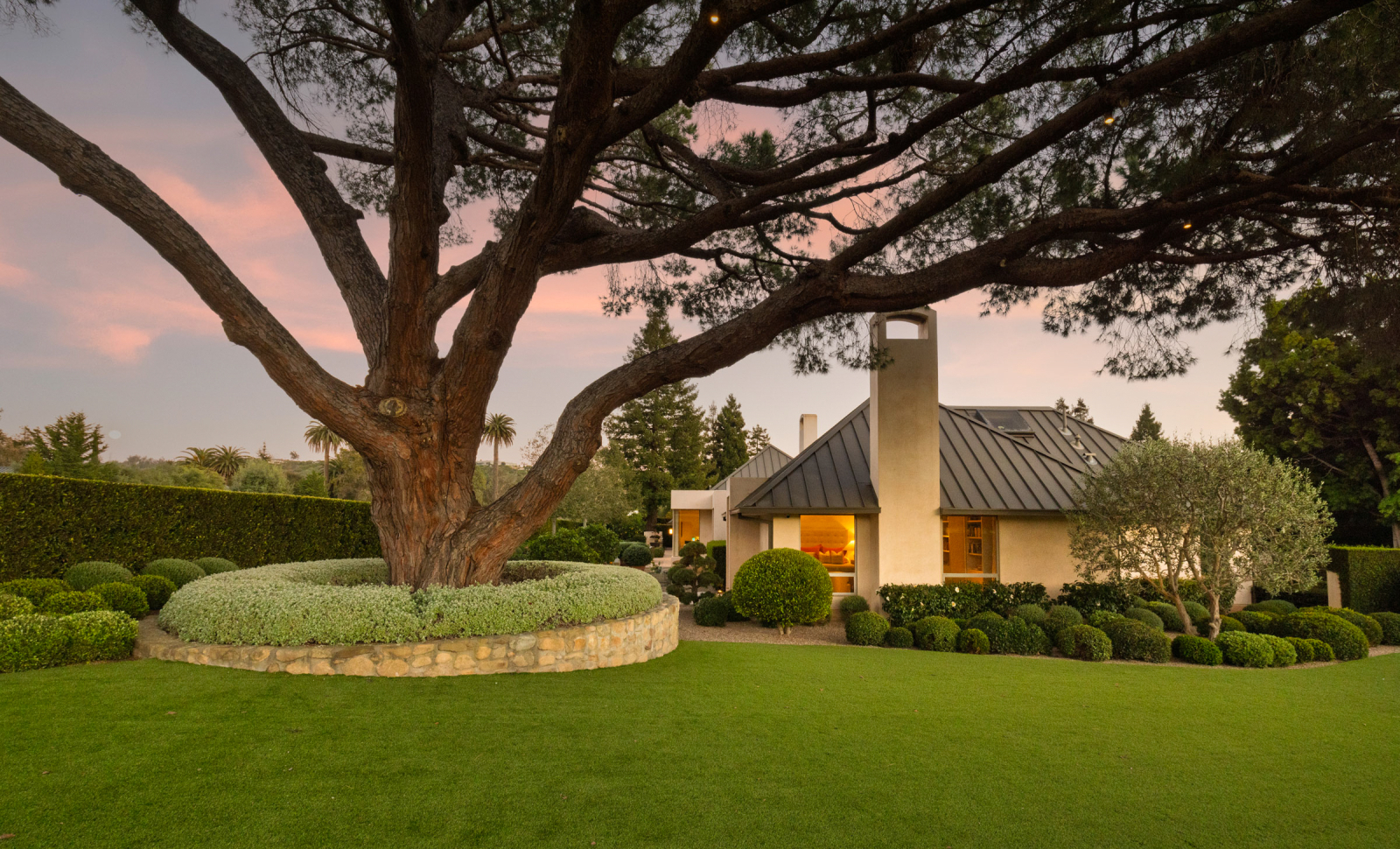 Elegant Montecito home at sunset framed by a massive mature tree, featuring manicured landscaping, glowing interior lights, and a peaceful garden setting.