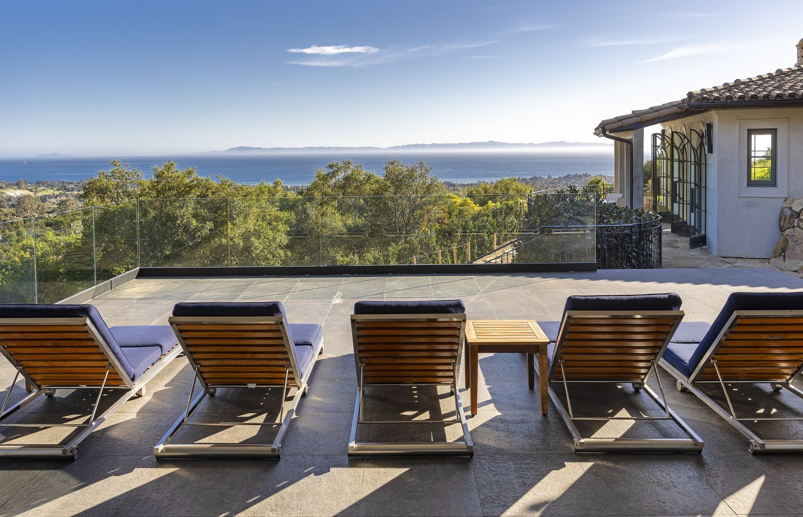 A row of lounge chairs lined up on a terrace, facing an ocean view over treetops.