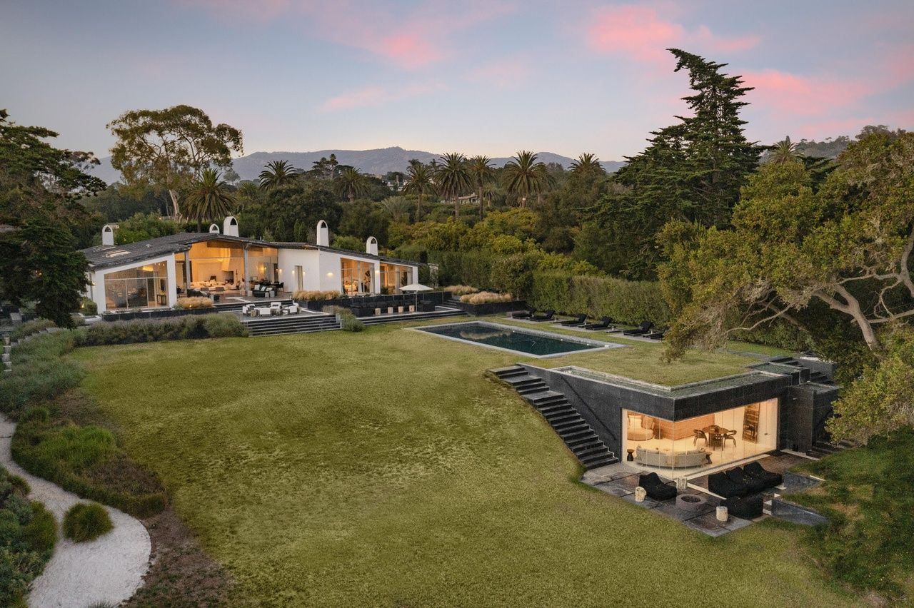 A stunning Wallace Neff designed home in Hope Ranch, Santa Barbara, with a separate guest huose and green lawn surrounded by foliage and a pink sunset