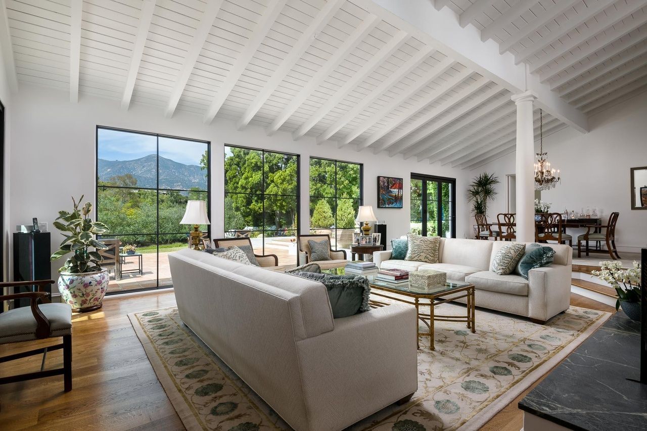 White, open-beam vaulted ceiling living room of a luxury home with stylish furnishings in front of floor to ceiling windows with a mountain view