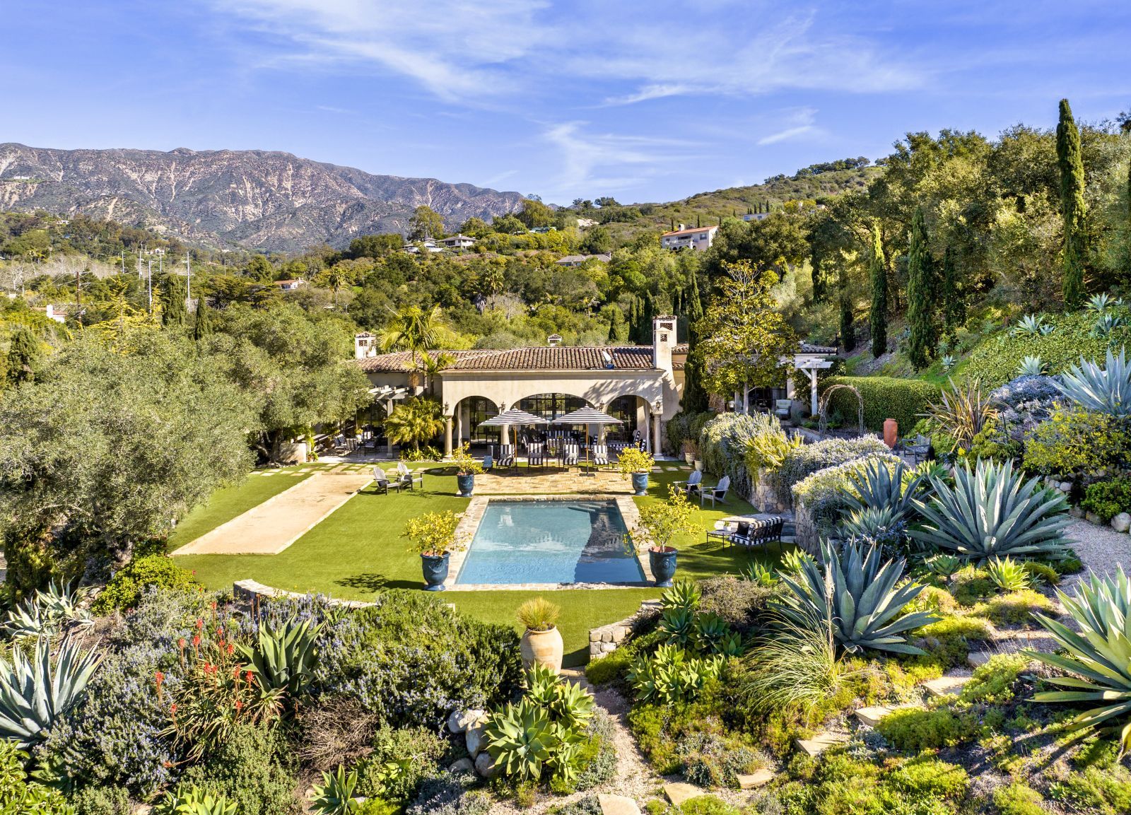 A Santa Barbara estate home surrounded by lush vegetation with a square pool in the foreground and mountains in the background.