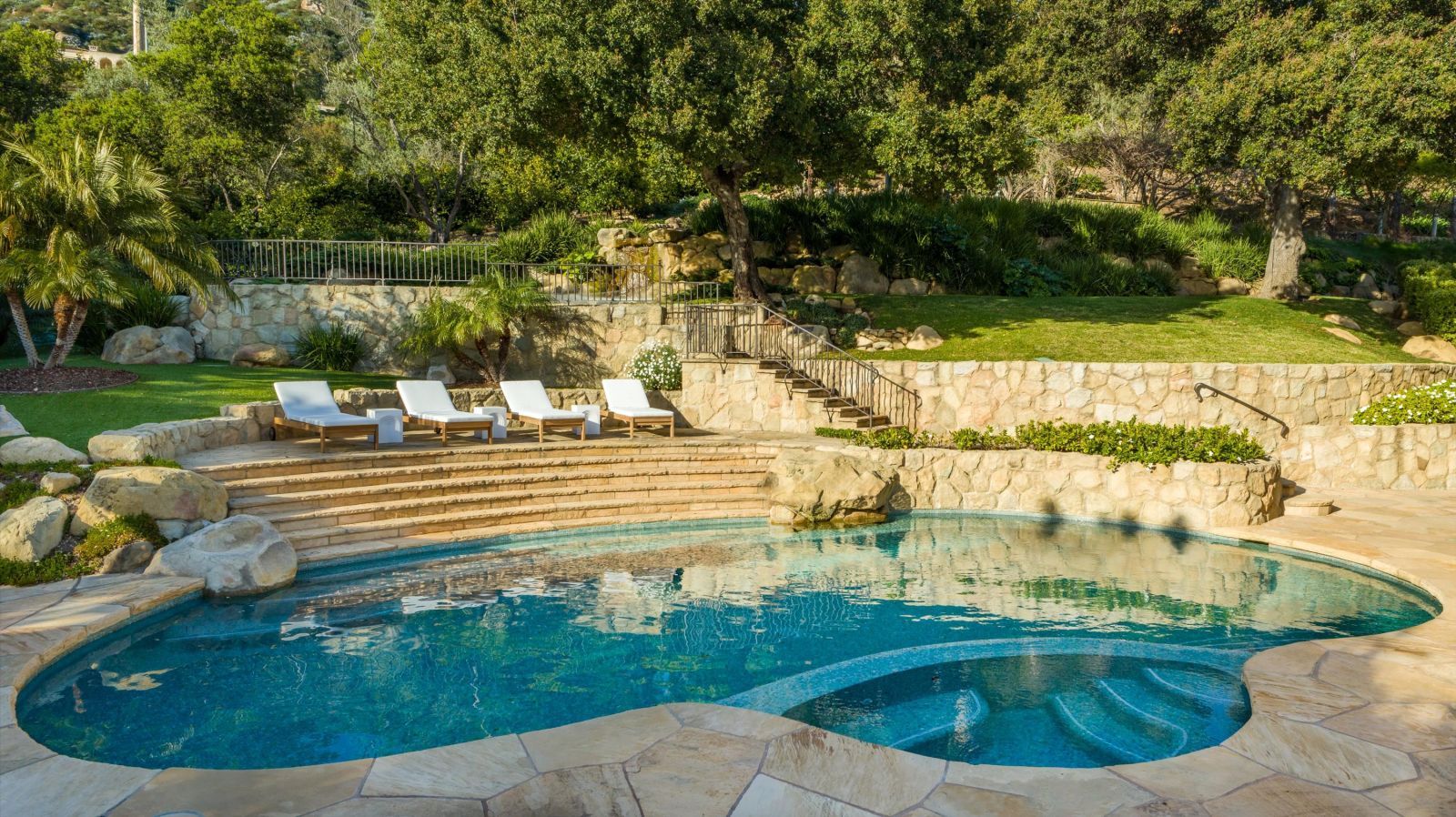 A luxury backyard pool area surrounded by lush vegetation and a row of lounge chairs at the top of stairs.