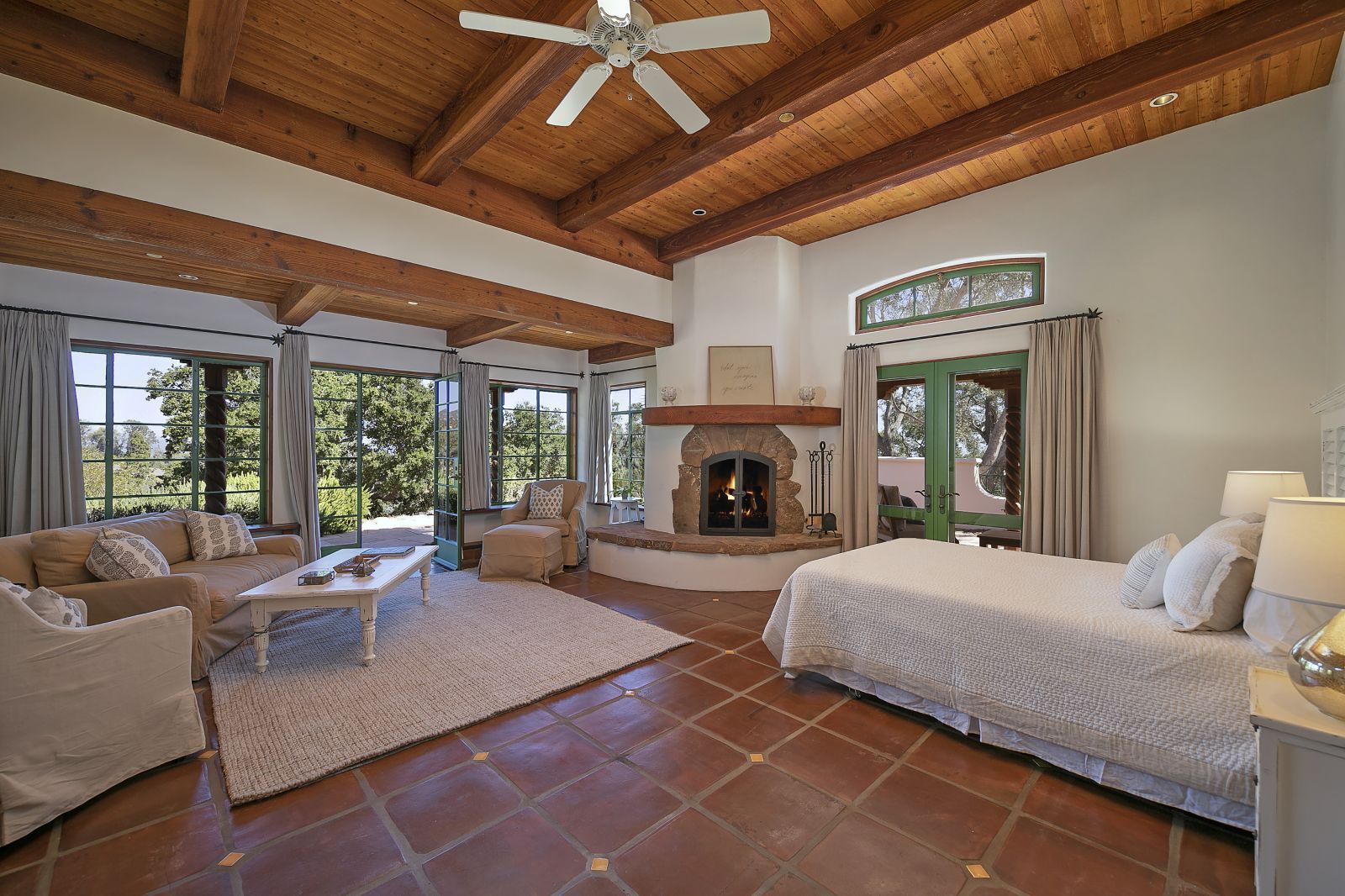 A spacious bedroom with terracotta tile floor, high wood-beam ceiling, a kiva fireplace, and multiple windows.