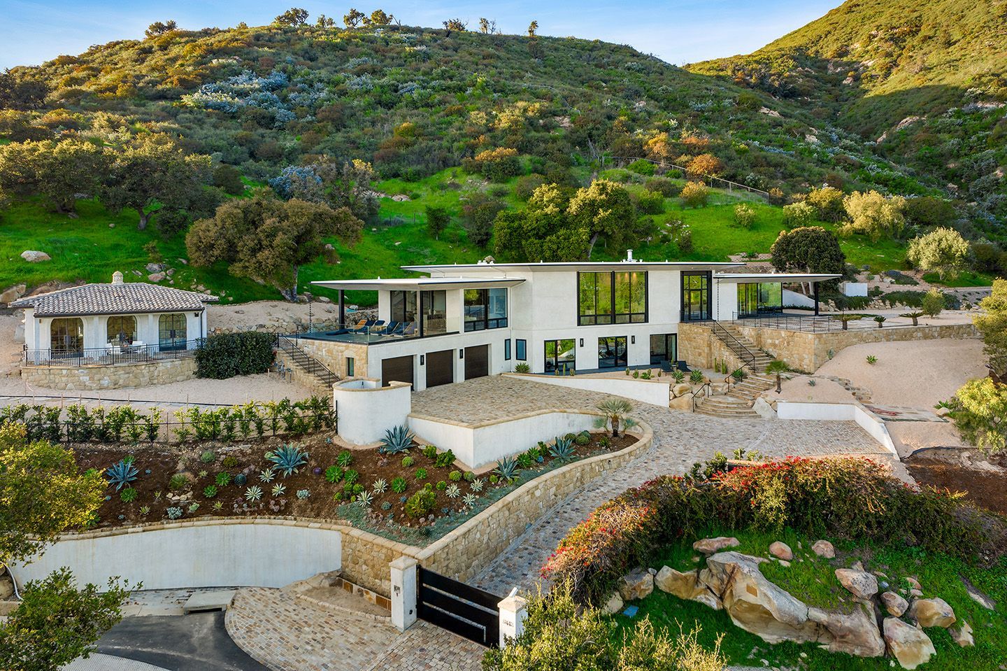 A contemporary Montecito estate, with a home and guest home with white walls, expanses of glass, and a verdant hillside in the background.