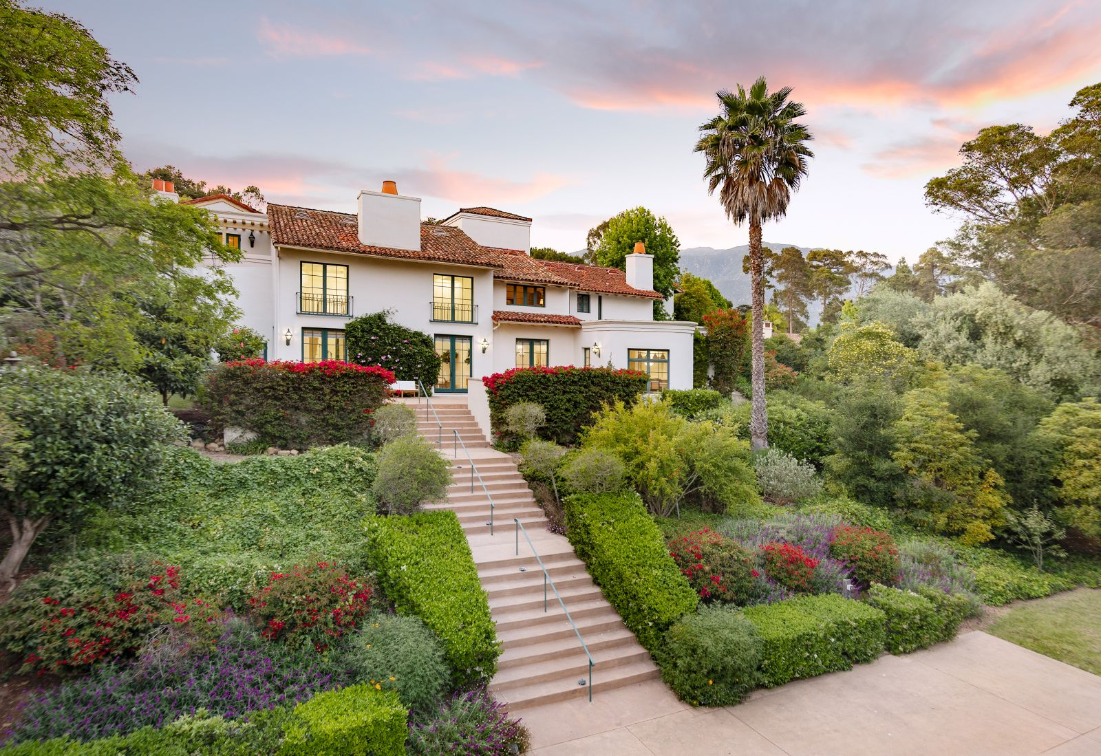 Montecito Spanish-style estate with white stucco walls, red tile roof, terraced landscaping, central staircase, and mountain backdrop at sunset.