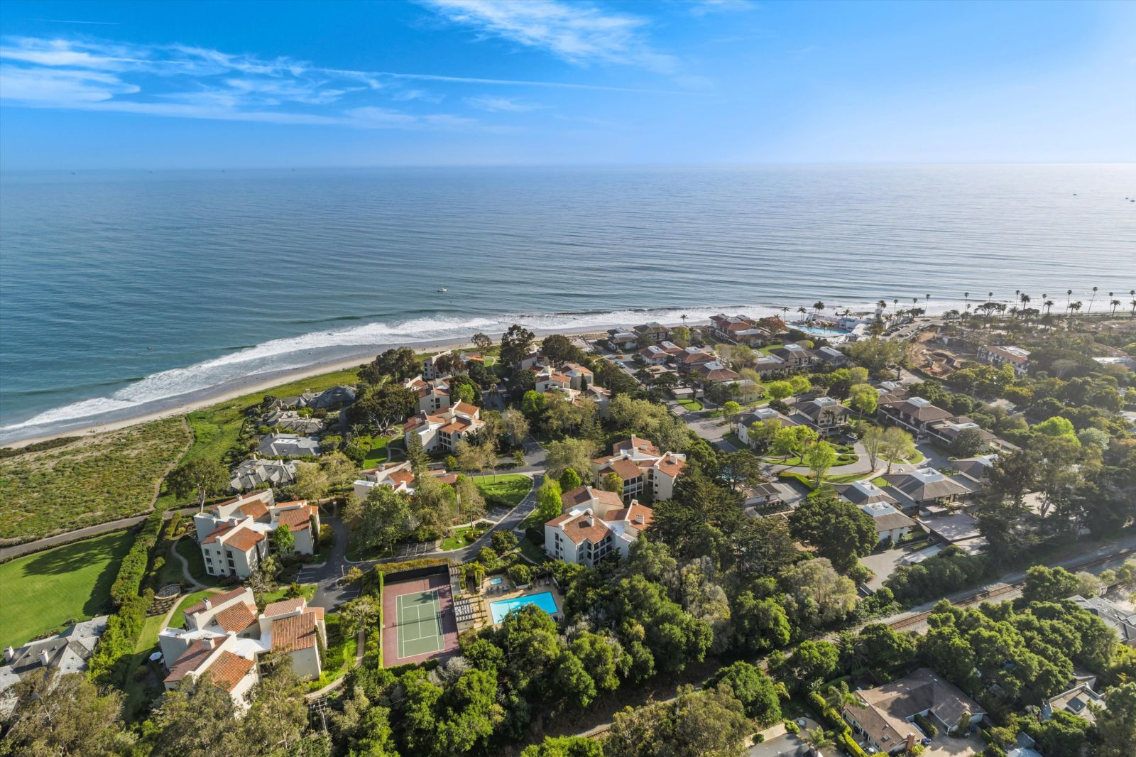 Aerial view of Montecito's coastline featuring luxury homes, tree-lined neighborhoods, tennis courts, and the Pacific Ocean stretching to the horizon under a clear blue sky.