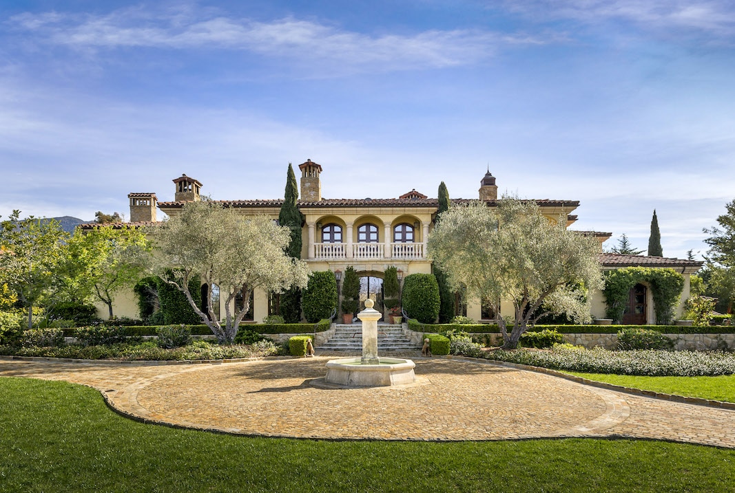 The front of a Montecito home that is currently for sale with a fountain in the foreground and some white fluffy clouds against a blue sky that naturally offers great air quality