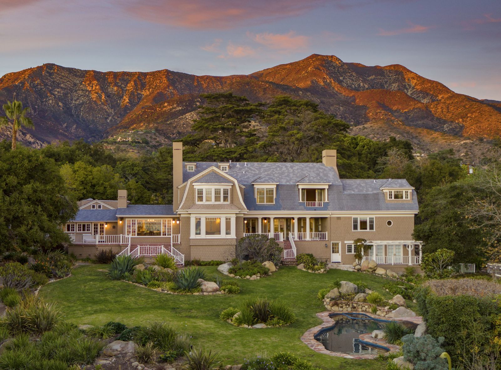 The exterior of a large Cape-Cod Style home in Montecito, with a sprawling lawn in front and mountains in the background glowing orange at duskns