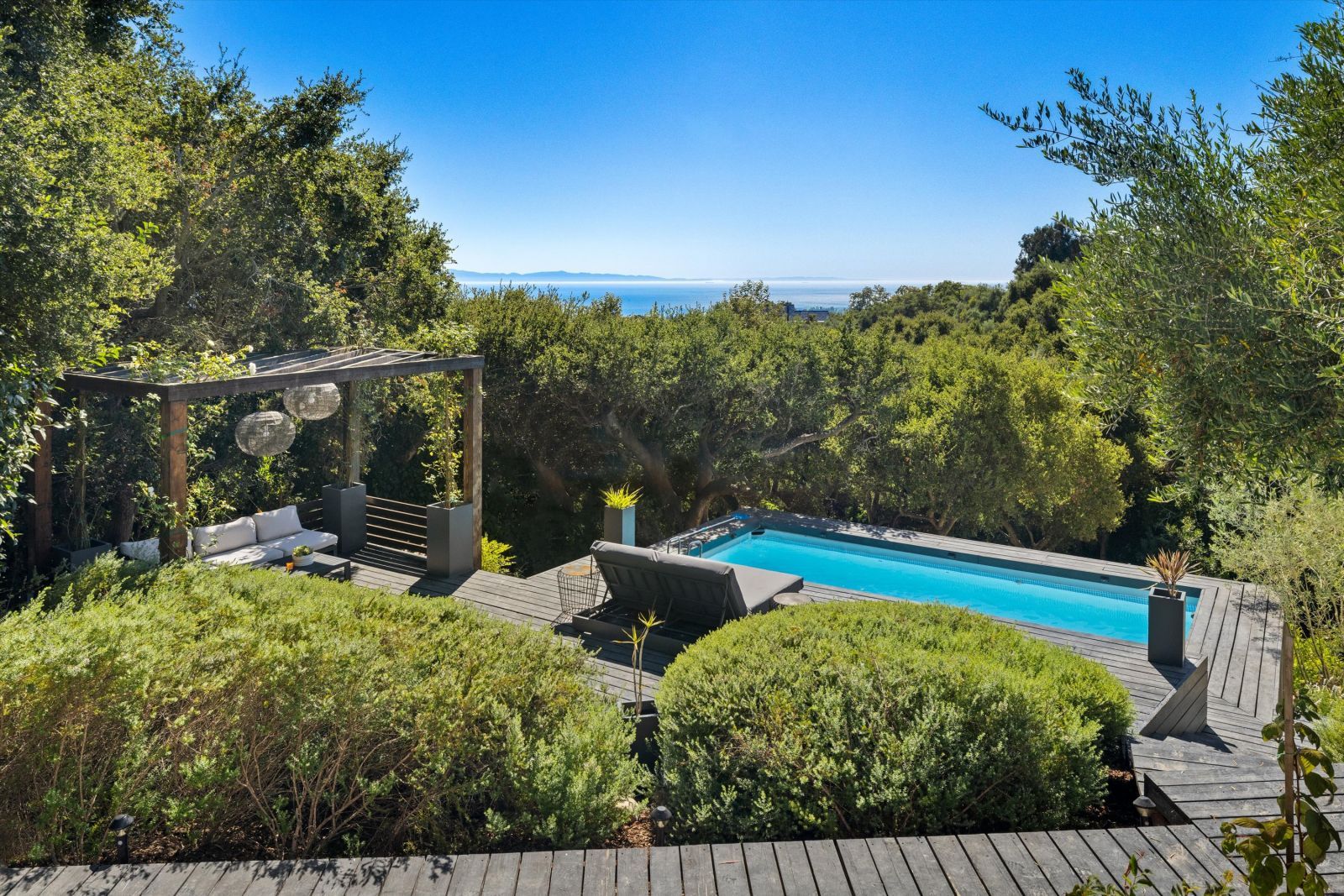 A sparkling blue pool with a double lounge chair on an expansive deck with a pergola, lush foliage, and an ocean view.