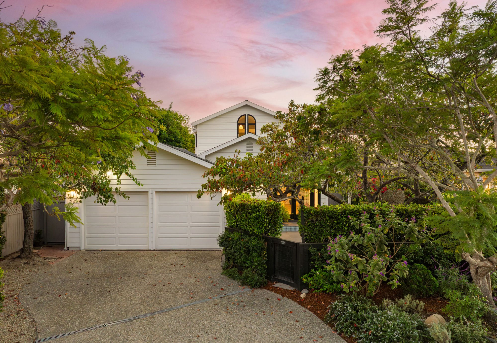 A charming Santa Barbara farmhouse–style home sits behind mature trees and lush landscaping at sunset. The residence features a three-car garage, inviting front entry, and a private garden setting, blending classic architecture with curb appeal in a peaceful residential neighborhood.