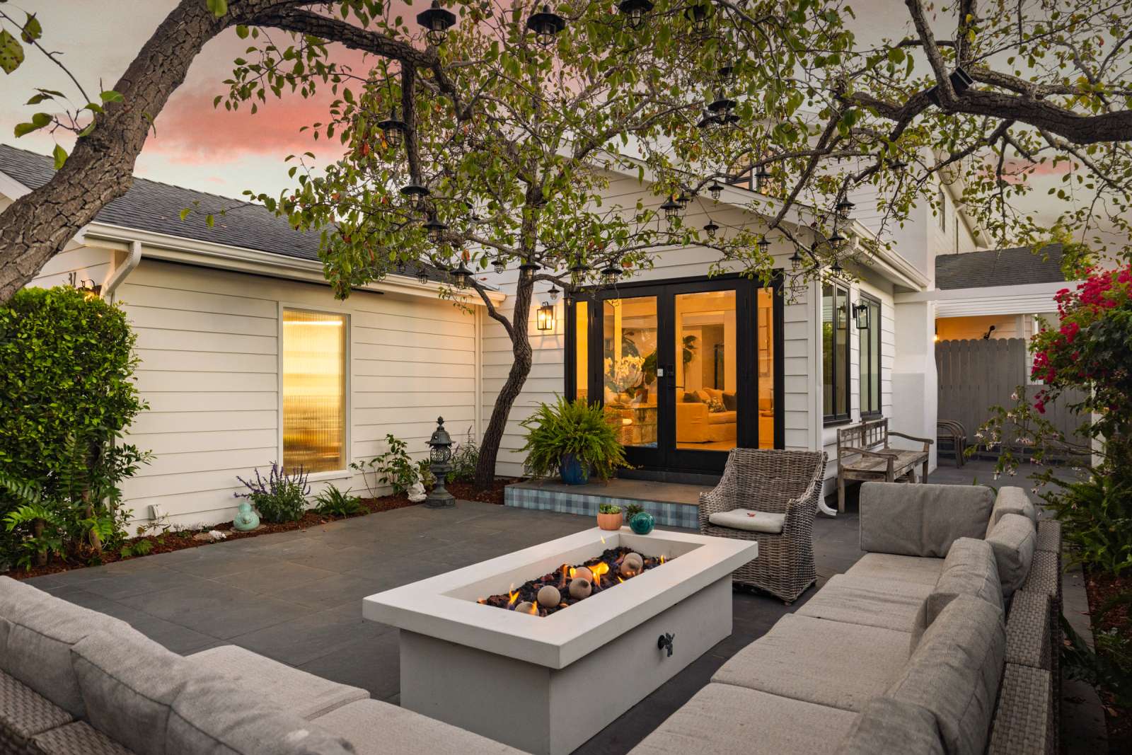 A beautifully designed outdoor living space features a modern fire table surrounded by comfortable seating beneath a mature tree strung with decorative lanterns. Large glass doors open to a warm, inviting interior, while lush landscaping and soft evening light create an intimate setting for year-round indoor-outdoor entertaining in Santa Barbara.