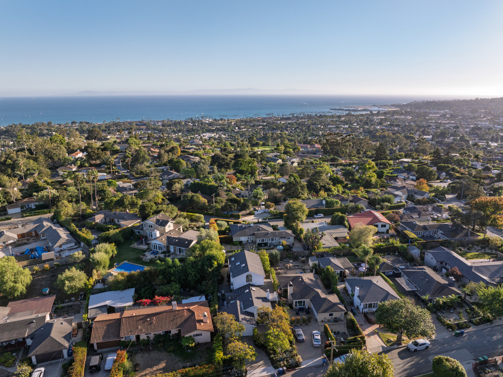 An aerial view of Eucalyptus Hill in Santa Barbara showcases a picturesque residential neighborhood nestled between the Santa Ynez Mountains and the Pacific Ocean. Tree-lined streets, charming homes, and lush landscaping stretch toward the coastline, highlighting the area's desirable location, ocean proximity, and scenic beauty.