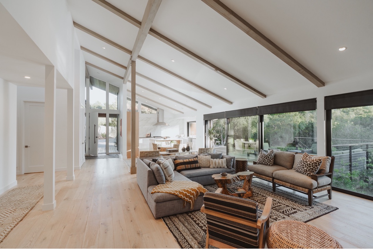 The living room of a home for sale in Montecito with high beamed ceilings and floor to ceiling windows