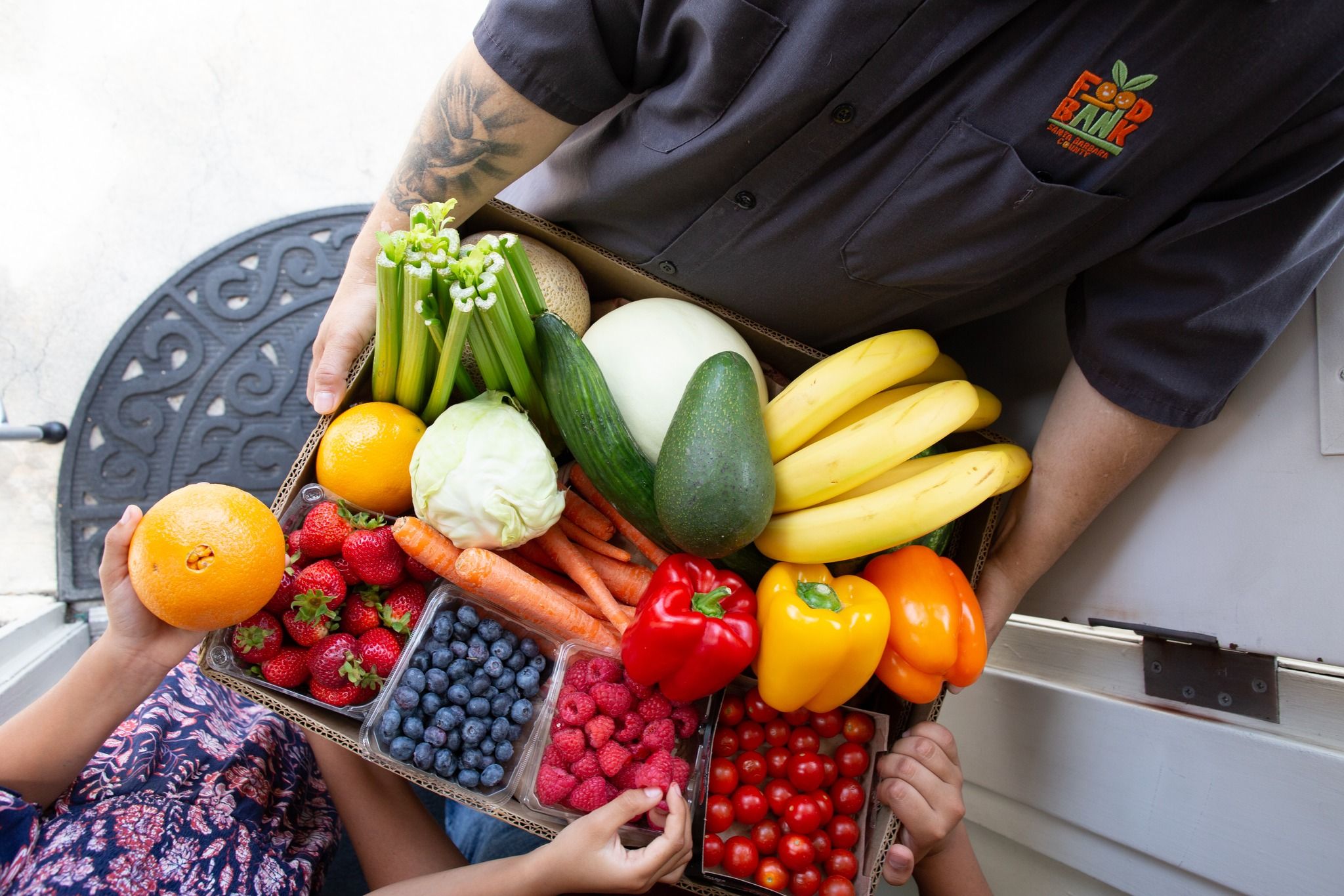 A man holding a basket of vibrantly colored  fruits and vegetables with a child picking an orange from the basket.