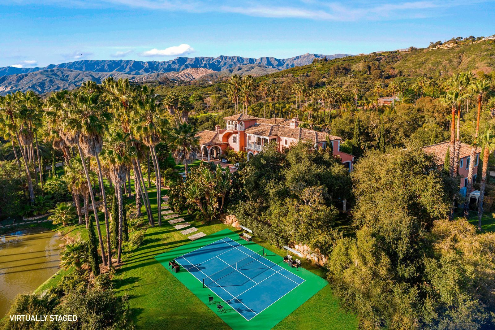 A birds eye view of a luxury home surrounded by green trees with a bright blue tennis court in the foreground surrounded by lush greenery.