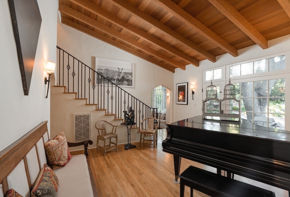 A grand piano sits in front of windows wiht trees in the background. there's also a staircase in the background.