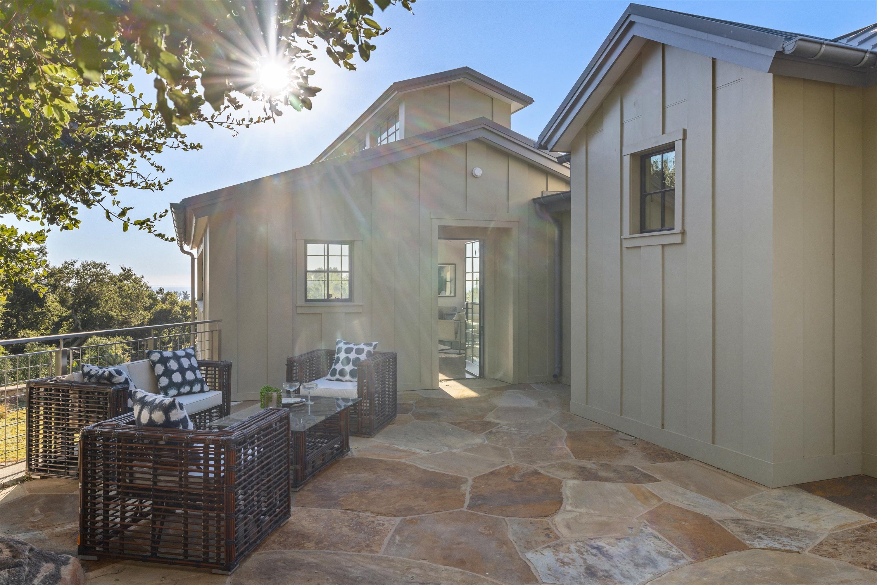 The outside of a Britt Jewett-designed farmhouse-style guest house, with its stone patio and upscale patio furniture in the foreground. The sun is casting golden rays through a tree's leaves