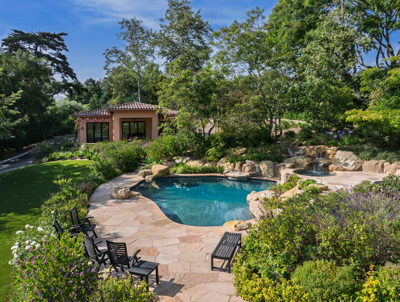 The backyard of a luxury home with a serene pool and red tile-roofed guest house, surrounded by lush vegetation.
