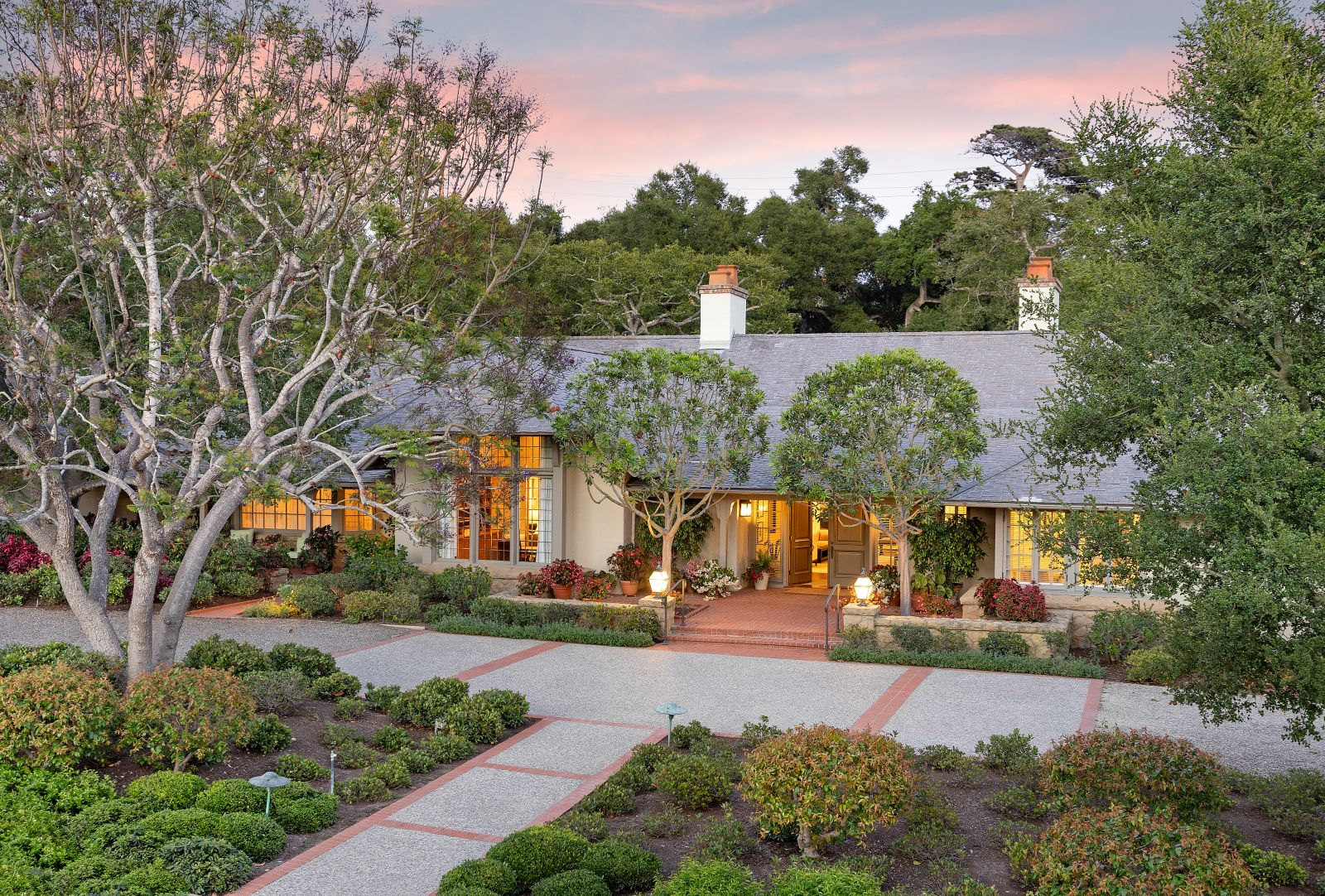 Charming single-story home at sunset with warm interior lights glowing through large windows, surrounded by mature trees, manicured landscaping, and a welcoming front entry with brick steps and garden-lined pathways.