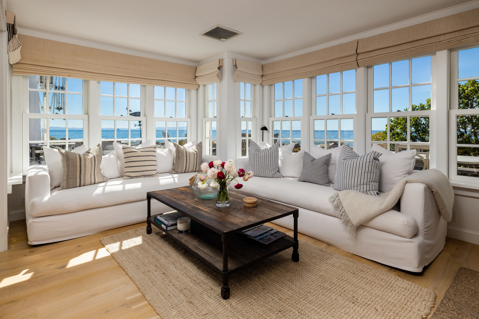 Bright coastal living room with white sectional sofas, striped pillows, wood coffee table, and expansive corner windows showcasing panoramic ocean views.