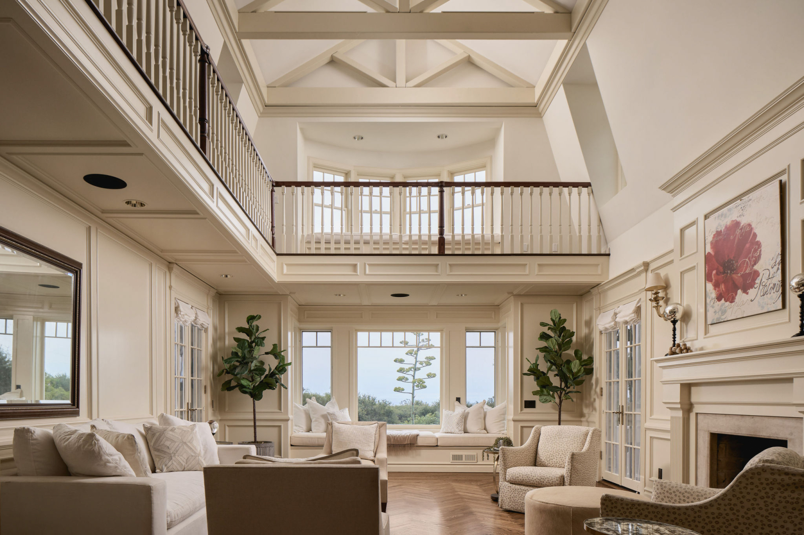 Bright, open-concept living room in a luxury Montecito home with vaulted ceilings, custom millwork, a fireplace, built-in window seating, and expansive windows overlooking lush greenery.