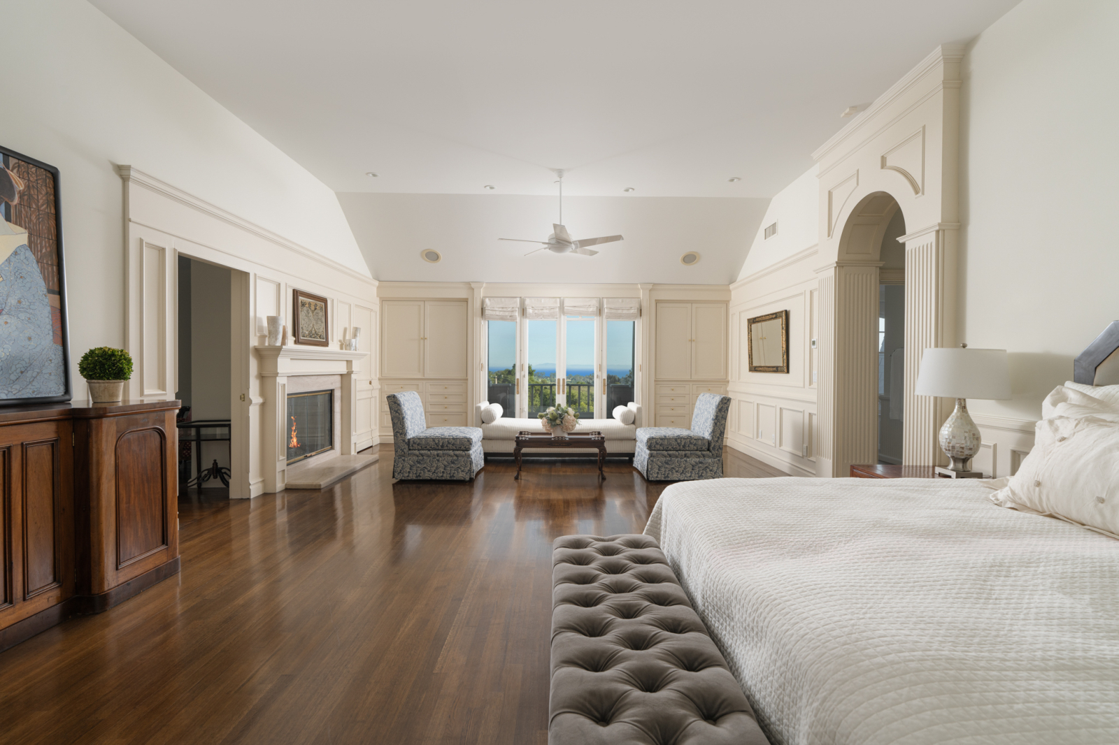 Spacious primary bedroom in a luxury Montecito home featuring hardwood floors, a fireplace, custom built-ins, a sitting area, and French doors opening to mountain and ocean views.