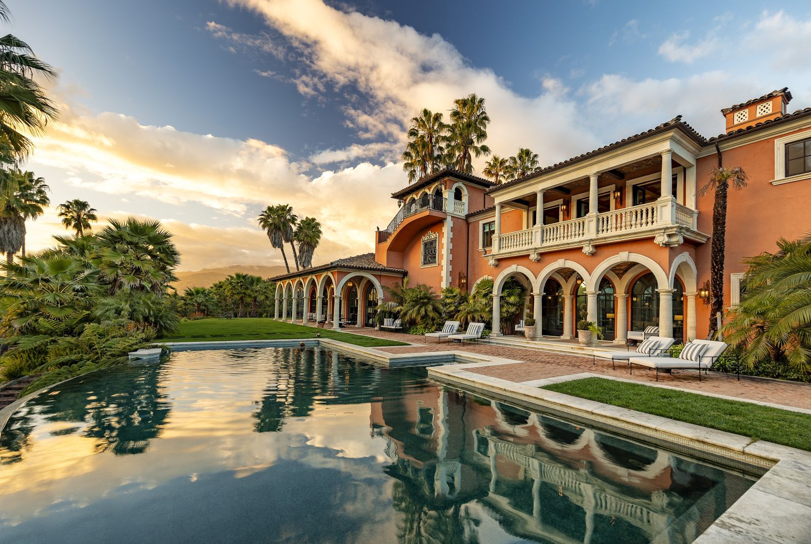 A world-class Hacienda-style Spanish Colonial estate with a serene pool in the foreground and fluffy clouds on a blue sky in the background.