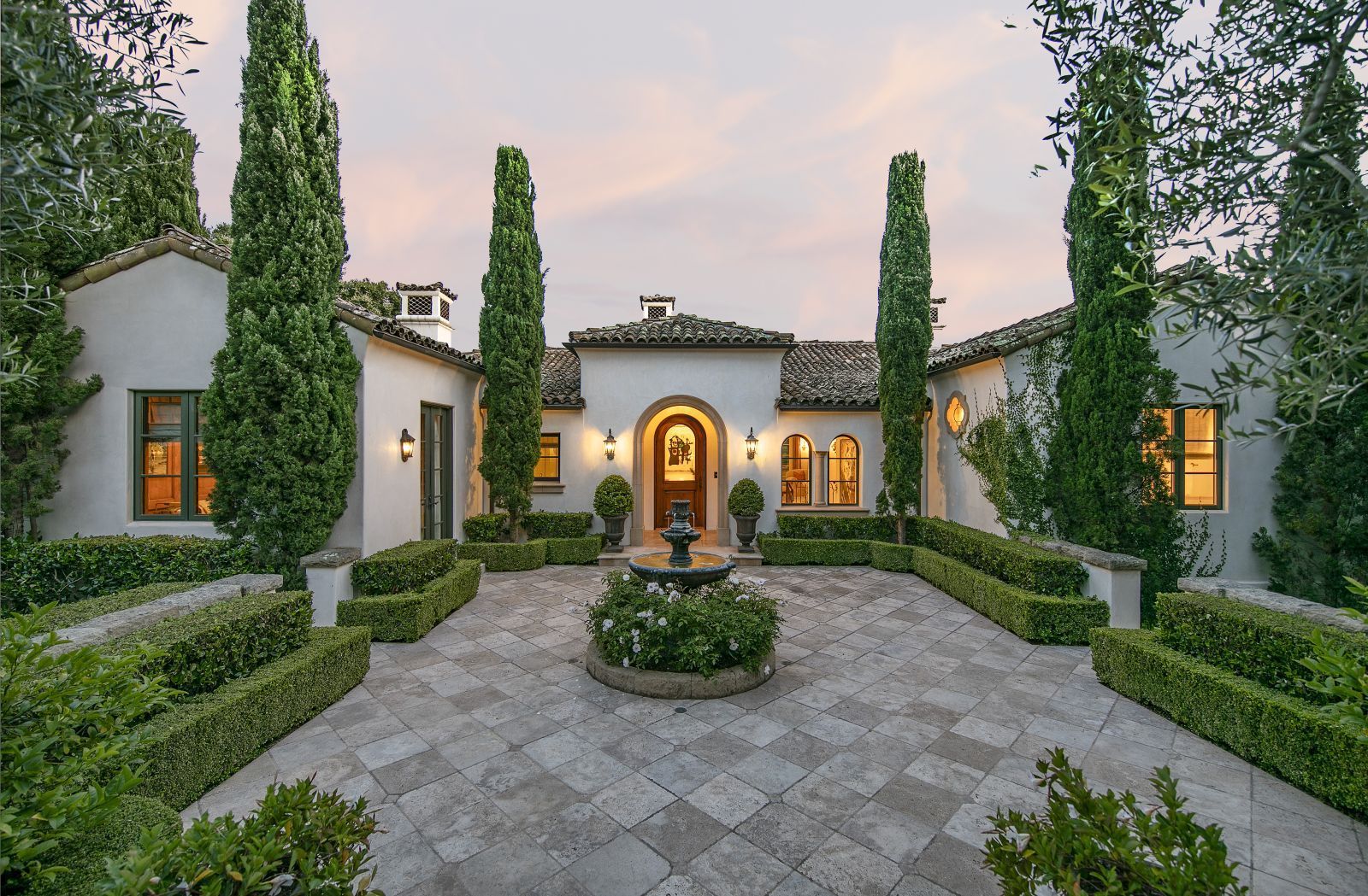 The meticulously manicured entry courtyard of a luxury home, with a border of privet hedges and Italian cypress trees. The home's facade has white walls and an arched front door, and is illuminated from within.