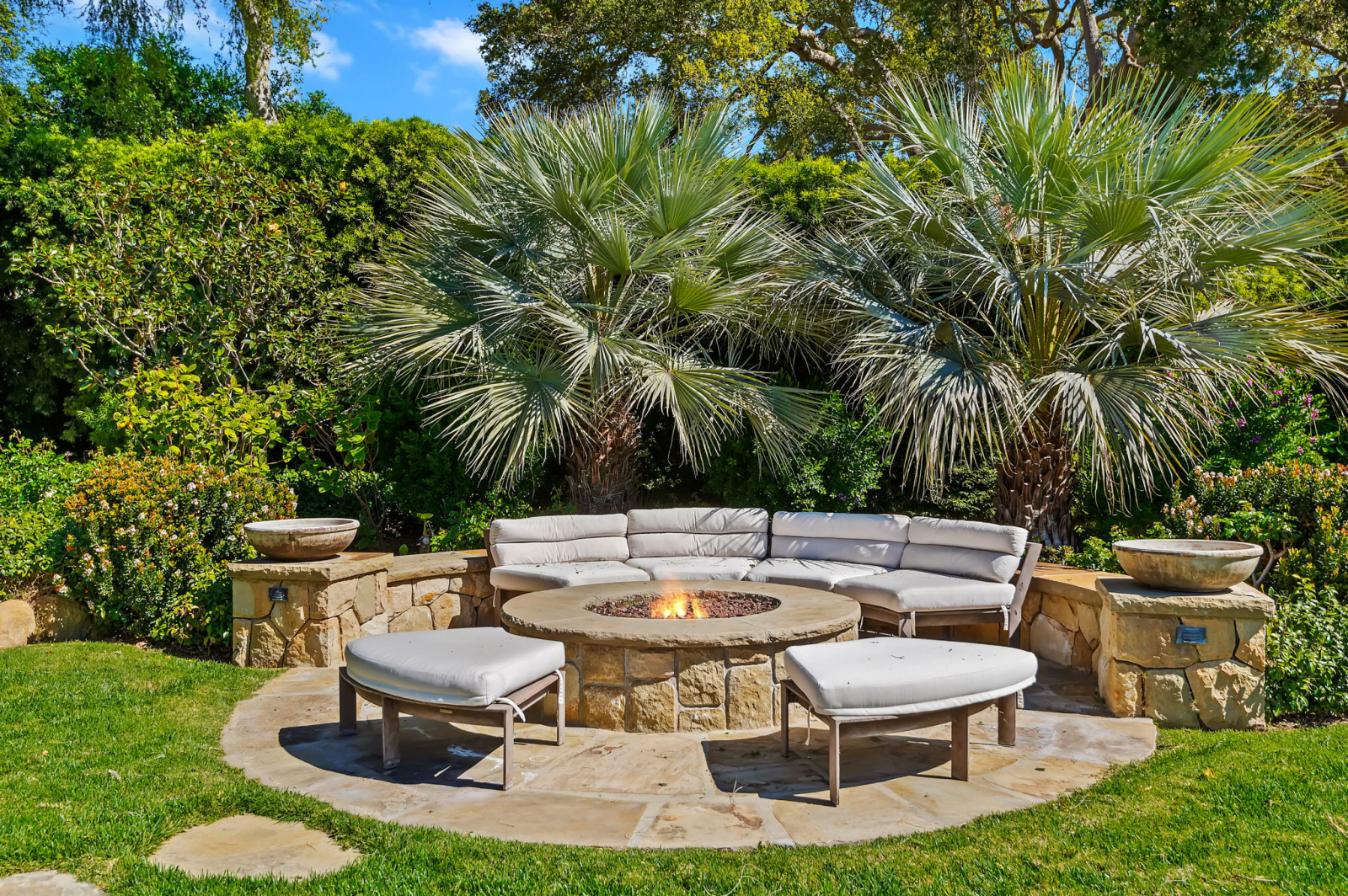 Luxury outdoor living area with a circular stone fire pit, cushioned seating, lush tropical landscaping, and manicured lawn, creating a private backyard retreat in a Santa Barbara estate.