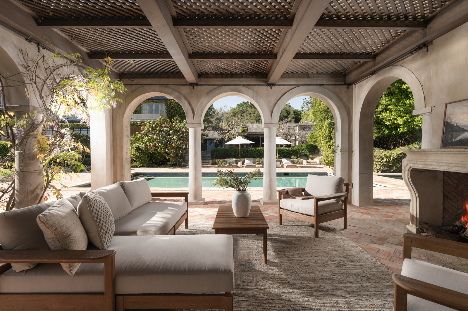 Covered outdoor patio with arched columns and wood-beamed ceiling, featuring cushioned seating and a fireplace, overlooking a swimming pool and landscaped garde