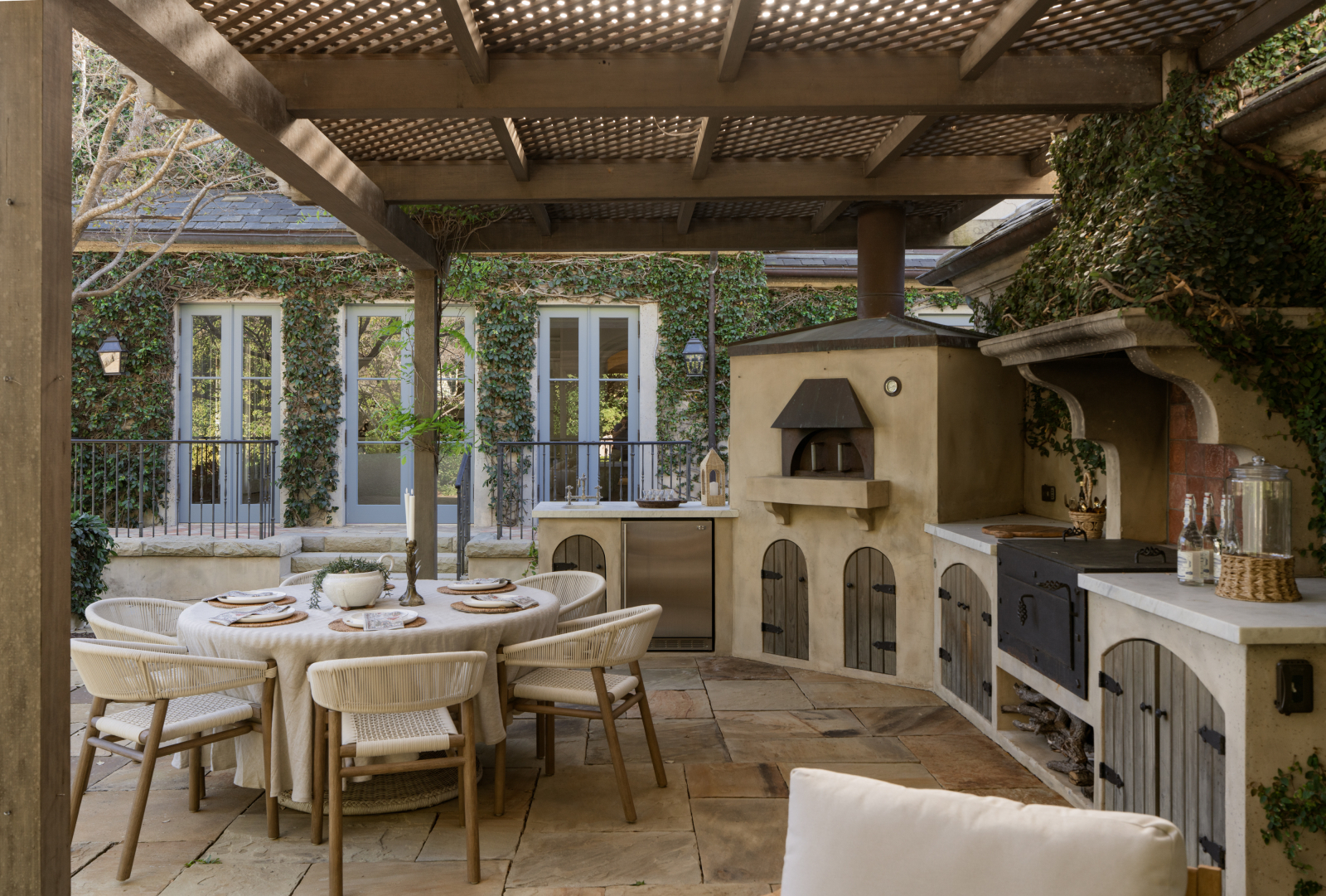 Luxury outdoor living space in Santa Barbara featuring a covered pergola, outdoor kitchen, pizza oven, dining area, and ivy-covered courtyard. The elegant entertaining area seamlessly blends indoor and outdoor living with high-end finishes, French doors, and resort-style amenities designed for year-round California living.