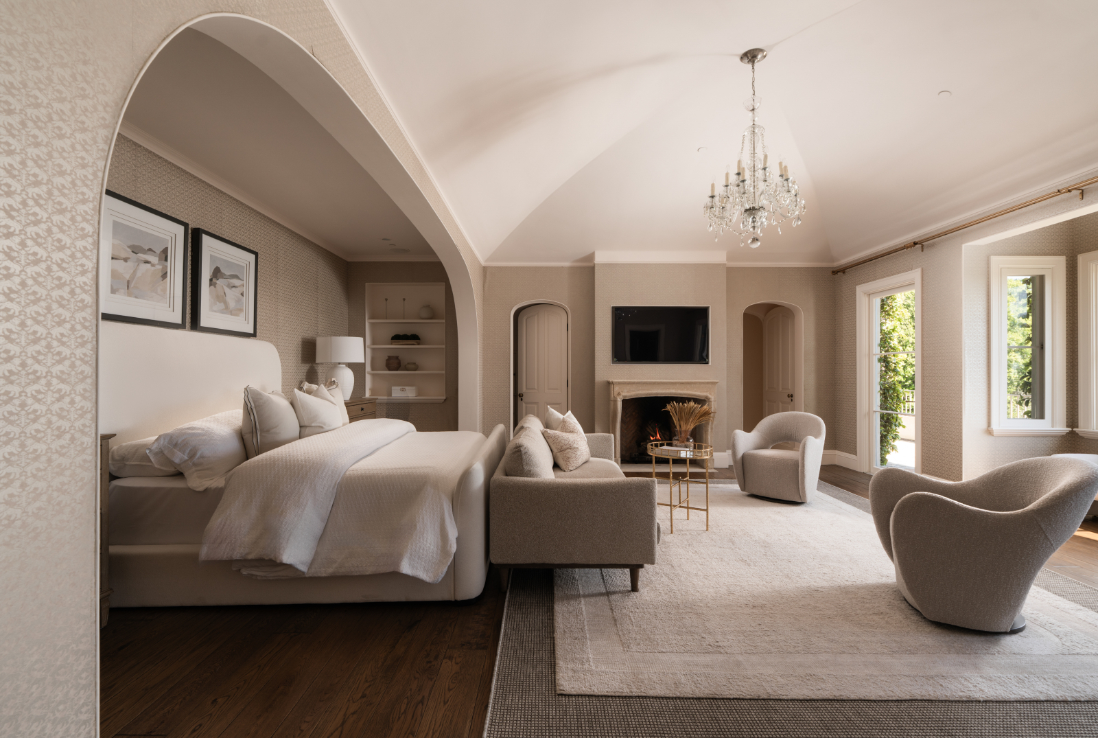 Elegant bedroom suite with a fireplace, sitting area, chandelier, and soft neutral furnishings, featuring arched architectural details and natural light from large windows.