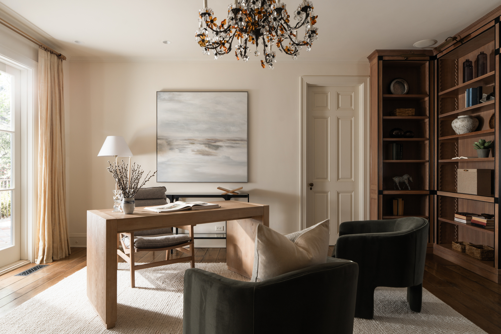 Elegant Montecito home office with a light wood desk, custom built-in bookshelves, hardwood floors, neutral décor, abstract artwork, and large windows providing abundant natural light.
