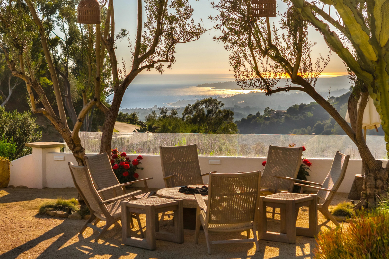 Luxury outdoor seating area with Adirondack-style chairs gathered around a fire pit, framed by mature olive trees and lush landscaping, overlooking the Pacific Ocean and the Santa Barbara coastline at sunset.