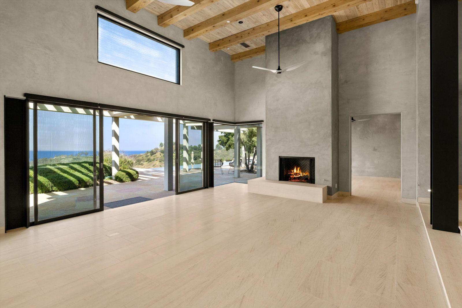An empty, expansive living room with its 17-foot plank and open beam vaulted ceiling, and fireplace, with doors open to a pergola-covered terrace.