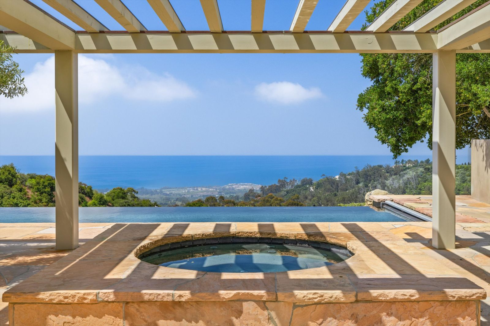 An in-ground hot tub under a pergola overlooking an infinity edge pool and the ocean.