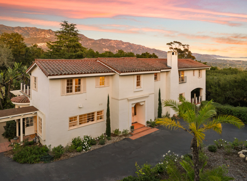 Montecito Spanish Colonial home with white stucco walls, red tile roof, arched architecture, landscaped grounds, and Santa Ynez Mountains at sunset.