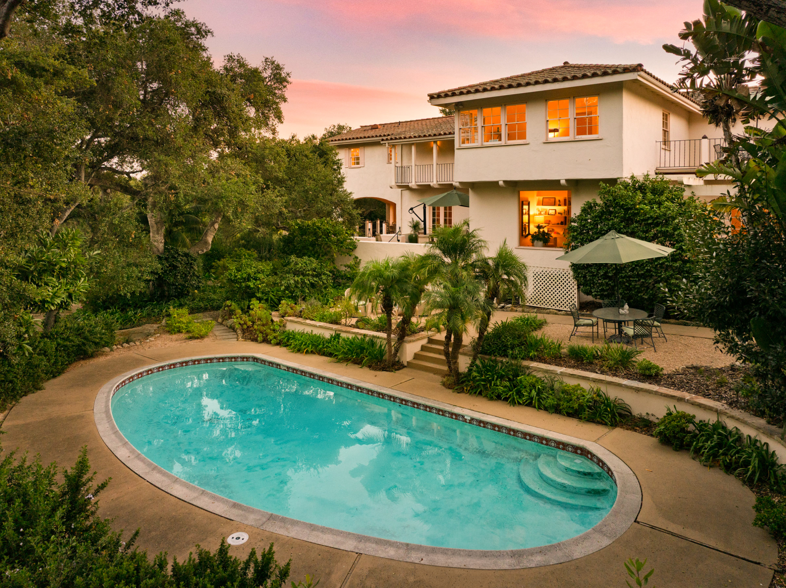 Spanish-style Montecito home overlooking a private swimming pool and lush tropical landscaping at sunset, with warm interior lighting and inviting outdoor living spaces.
