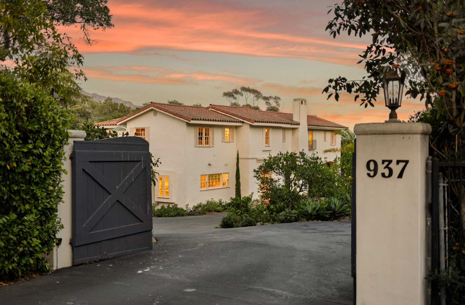 Spanish-style home with a red tile roof and gated driveway at sunset, surrounded by lush landscaping and warm interior lighting.